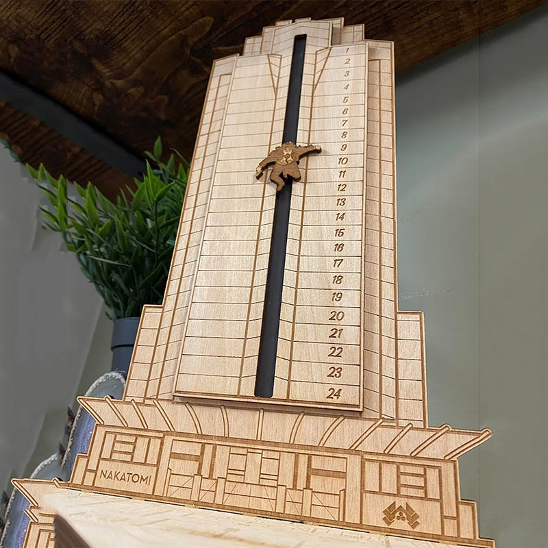 "Detaljerad trä Adventskalender med Die Hard-motiv, visar Hans Gruber faller från brunblå Nakatomi Plaza, roterbar design."